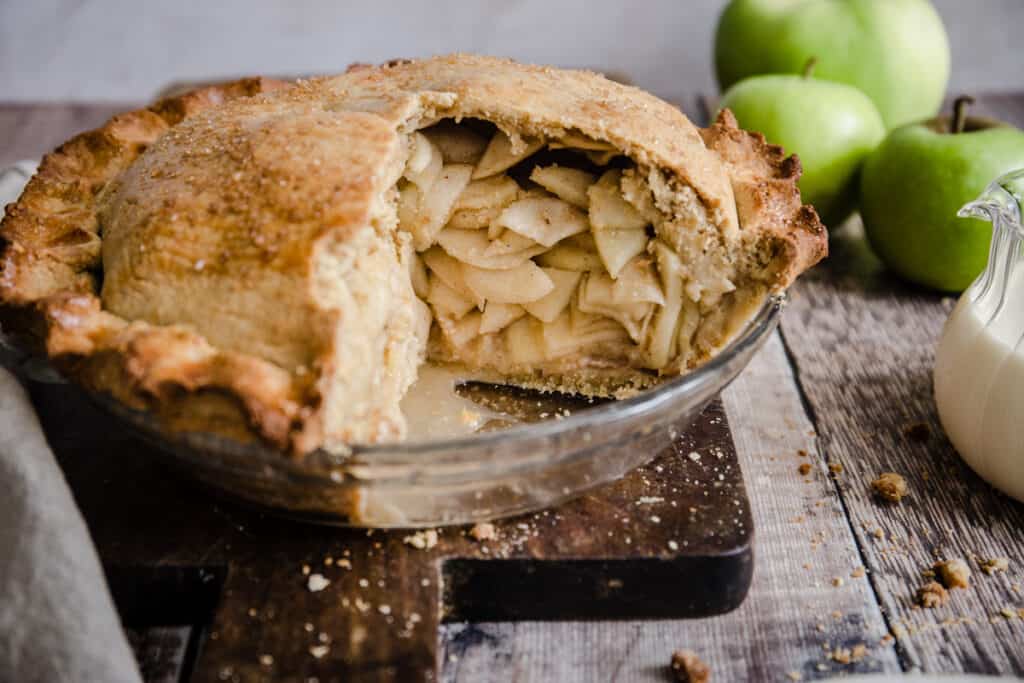 Cut open Apple pie on a wooden board next to apples and cream