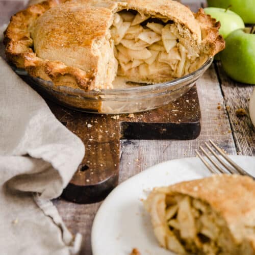 Cut open apple pie on a wooden board next to apples