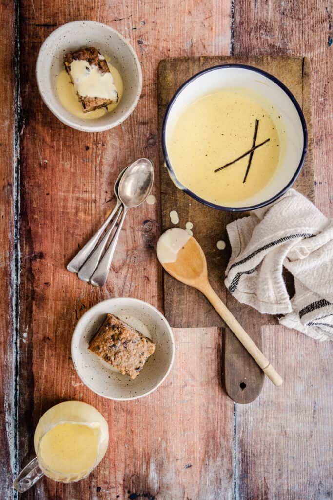custard in saucepan next to wooden spoon, bowls of dessert, spoons and jug of custard