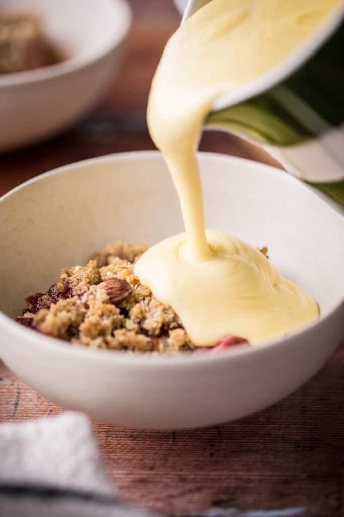 custard in saucepan next to wooden spoon, bowls of dessert, spoons and jug of custard