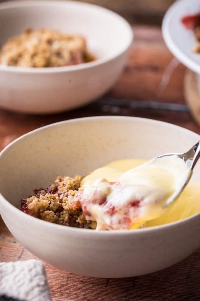 custard pouring from jug into a bowl of dessert next to spoons