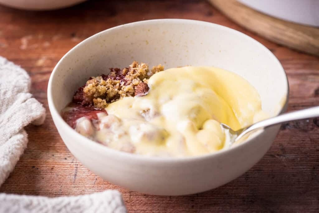 custard in saucepan next to wooden spoon, bowls of dessert, spoons and jug of custard