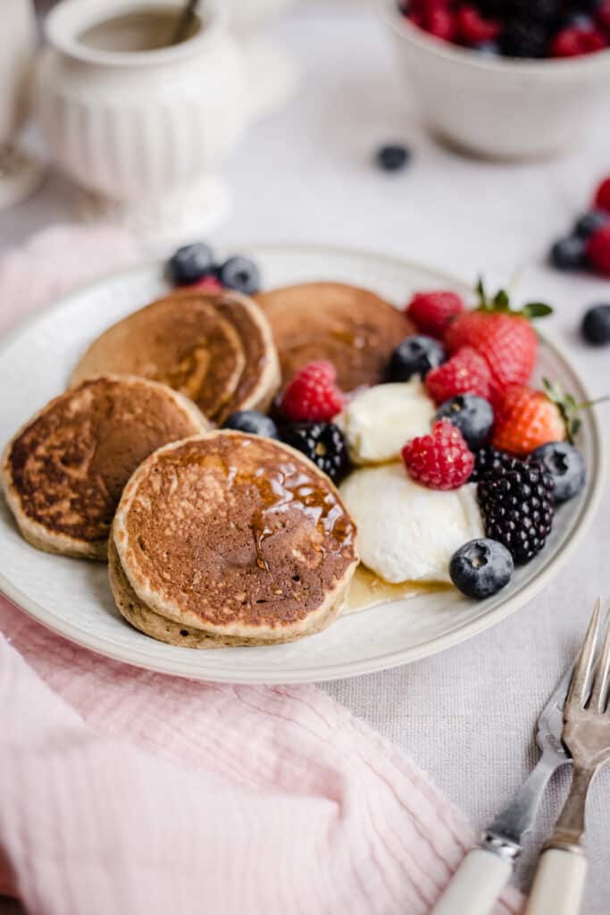 Pancakes on a plate with berries next to bowl of fruit, forks and honey pot