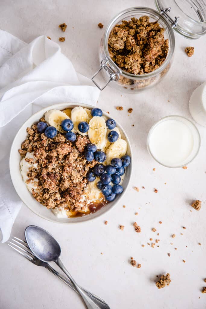 bowl of granola with blueberries, bananas next to spoon, milk and linen