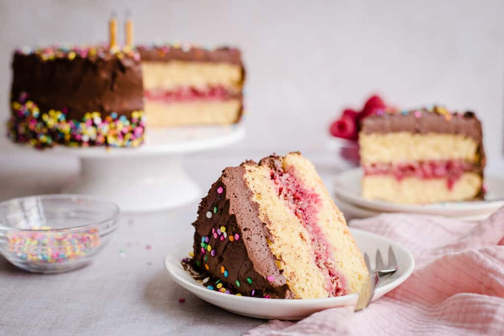 slice of cake in front of whole birthday cake and bowls of raspberries and sprinkles around
