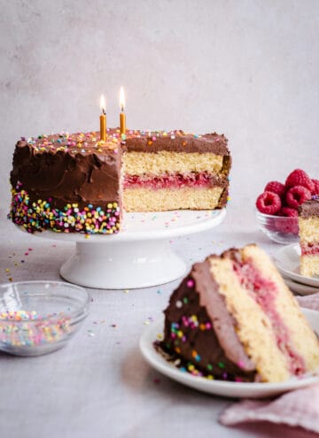 Birthday cake on a stand with slice of cake in front and bowls of raspberries and sprinkles around