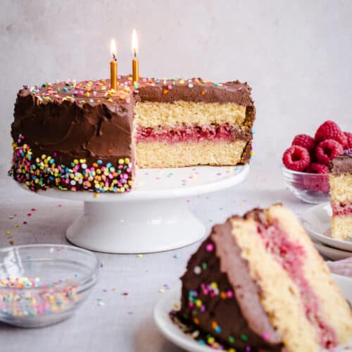 Birthday cake on a stand with slice of cake in front and bowls of raspberries and sprinkles around