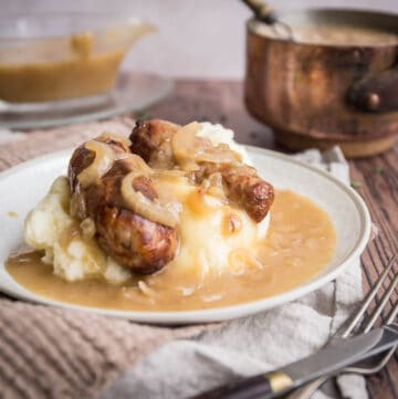 onion gravy over a plate of sausage and mash next to cutlery, gravy boat and saucepan