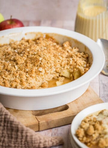 Gluten Free Apple Crumble in serving dish in front of apple, jug of custard, serving spoon and bowl of crumble