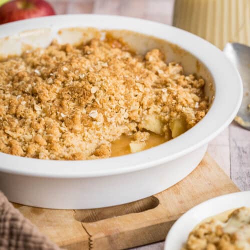 Gluten Free Apple Crumble in serving dish in front of apple, jug of custard, serving spoon and bowl of crumble
