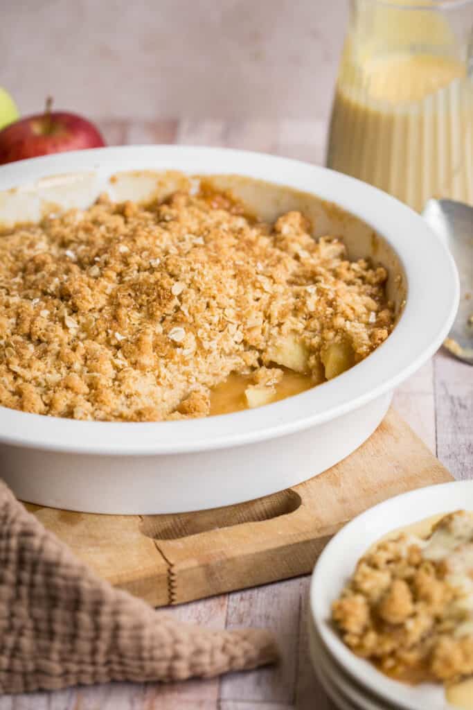 Gluten Free Apple Crumble in serving dish in front of apple, jug of custard, serving spoon and bowl of crumble