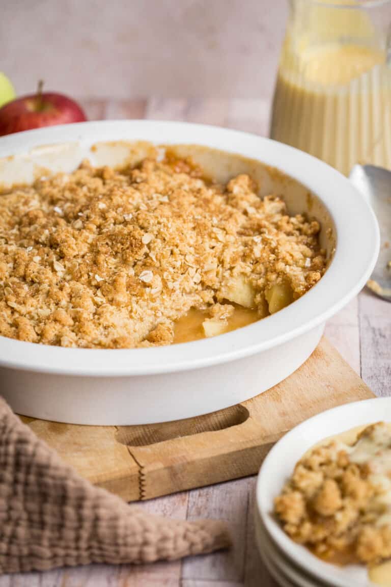 Gluten Free Apple Crumble in serving dish in front of apple, jug of custard, serving spoon and bowl of crumble