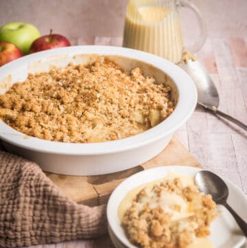 Gluten Free Apple Crumble in serving dish in front of apple, jug of custard, serving spoon and bowl of crumble
