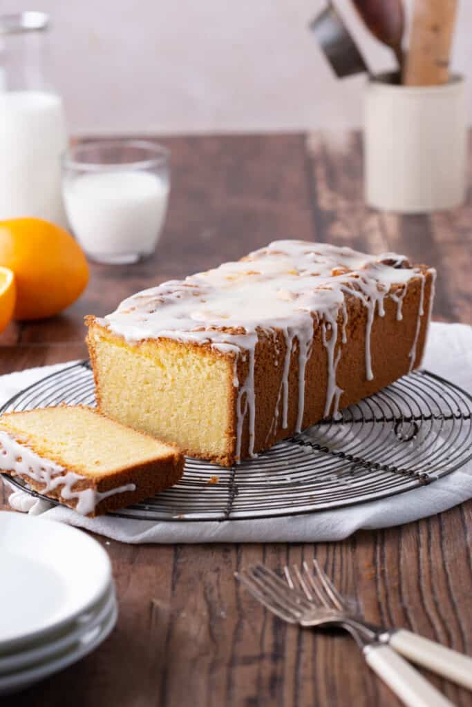 Gluten-Free Orange Drizzle Cake cut on a wire rack next to plates and oranges.