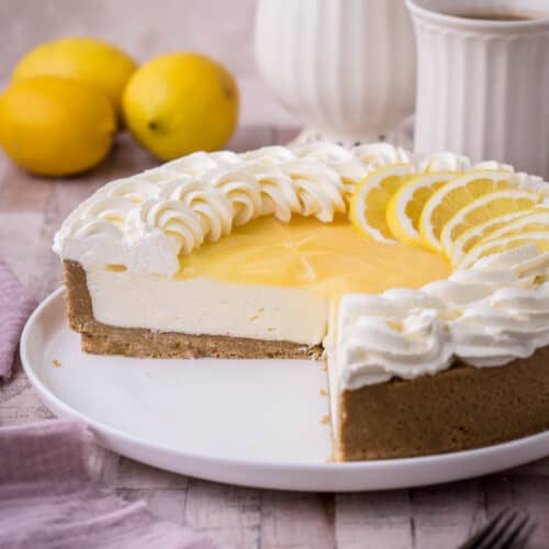 Lemon Curd Cheesecake on a plate. Decorated with lemon curd topping, whipped cream and fresh lemon slices. With large slice removed showing firm creamy centre.