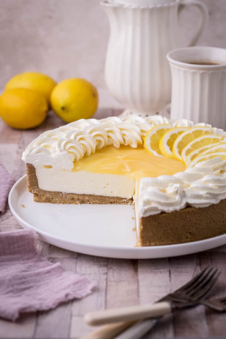 Lemon Curd Cheesecake on a plate. Decorated with lemon curd topping, whipped cream and fresh lemon slices. With large slice removed showing firm creamy centre.