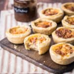 Mini Quiche on a wooden board. One with bite taken out showing meaty creamy inside.