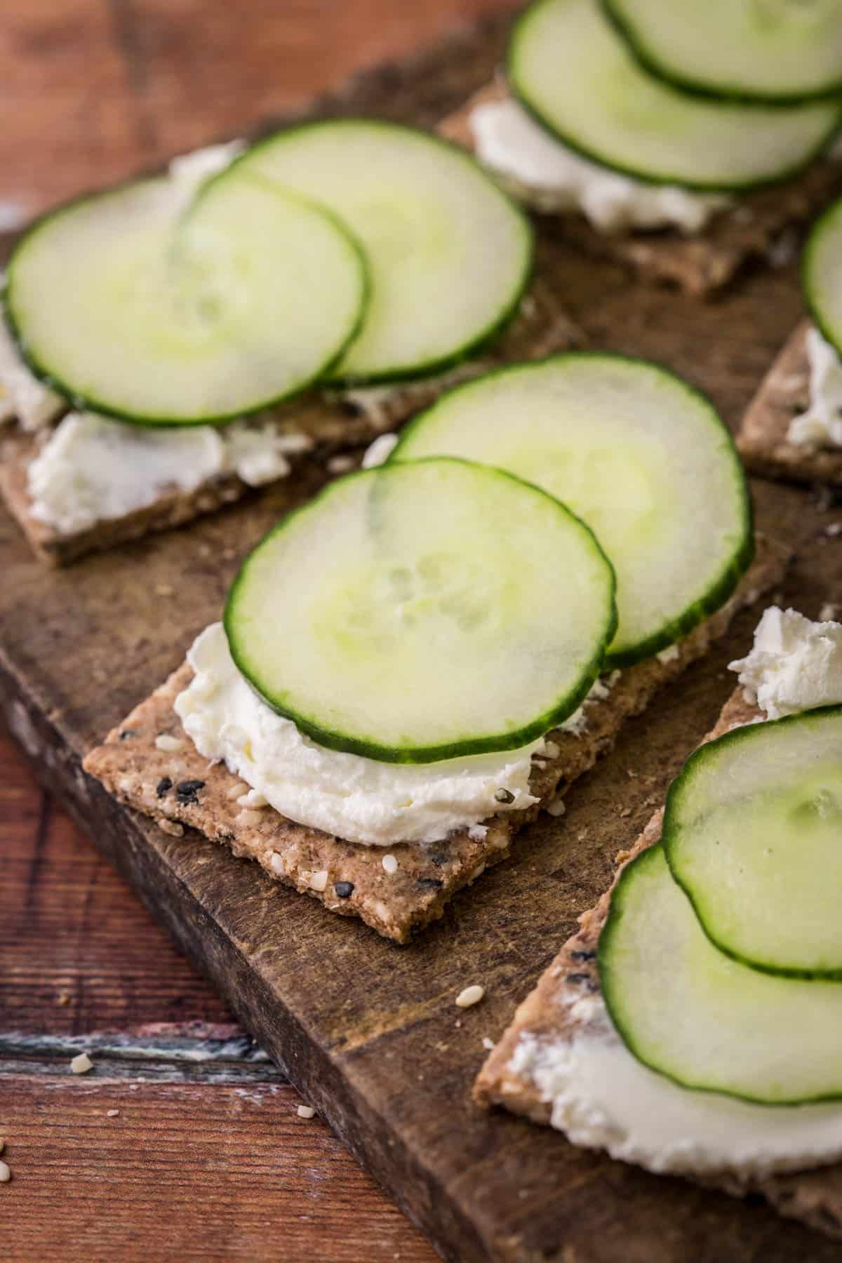 Seeded Gluten-Free Crispbreads spread with cream cheese and cucumber on wooden board.