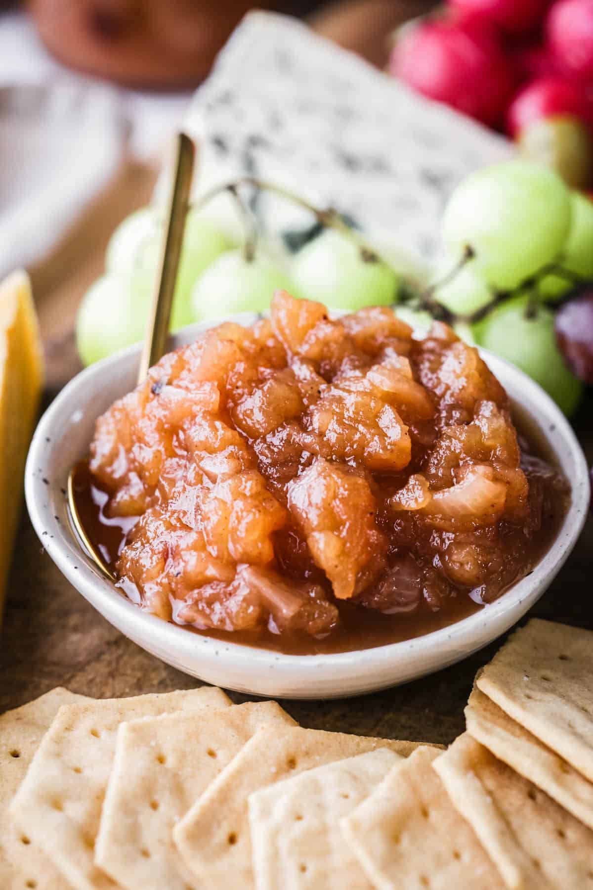 Bowl of Apple Ginger Chutney on a board surrounded by crackers, cheese, charcuterie, grapes.
