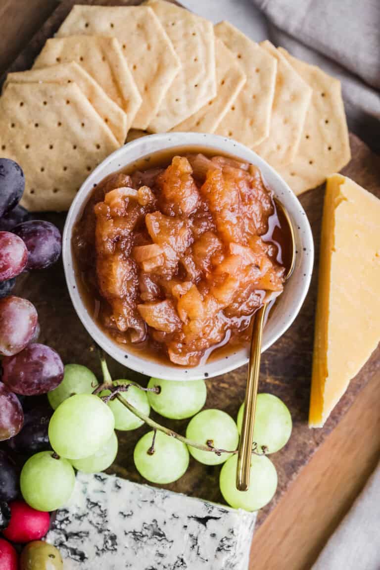 Bowl of Apple Ginger Chutney on a board surrounded by crackers, cheese, charcuterie, grapes.
