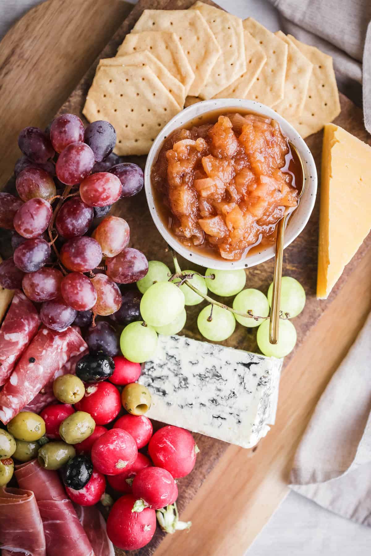 Bowl of Apple Ginger Chutney on a board surrounded by crackers, cheese, charcuterie, grapes.