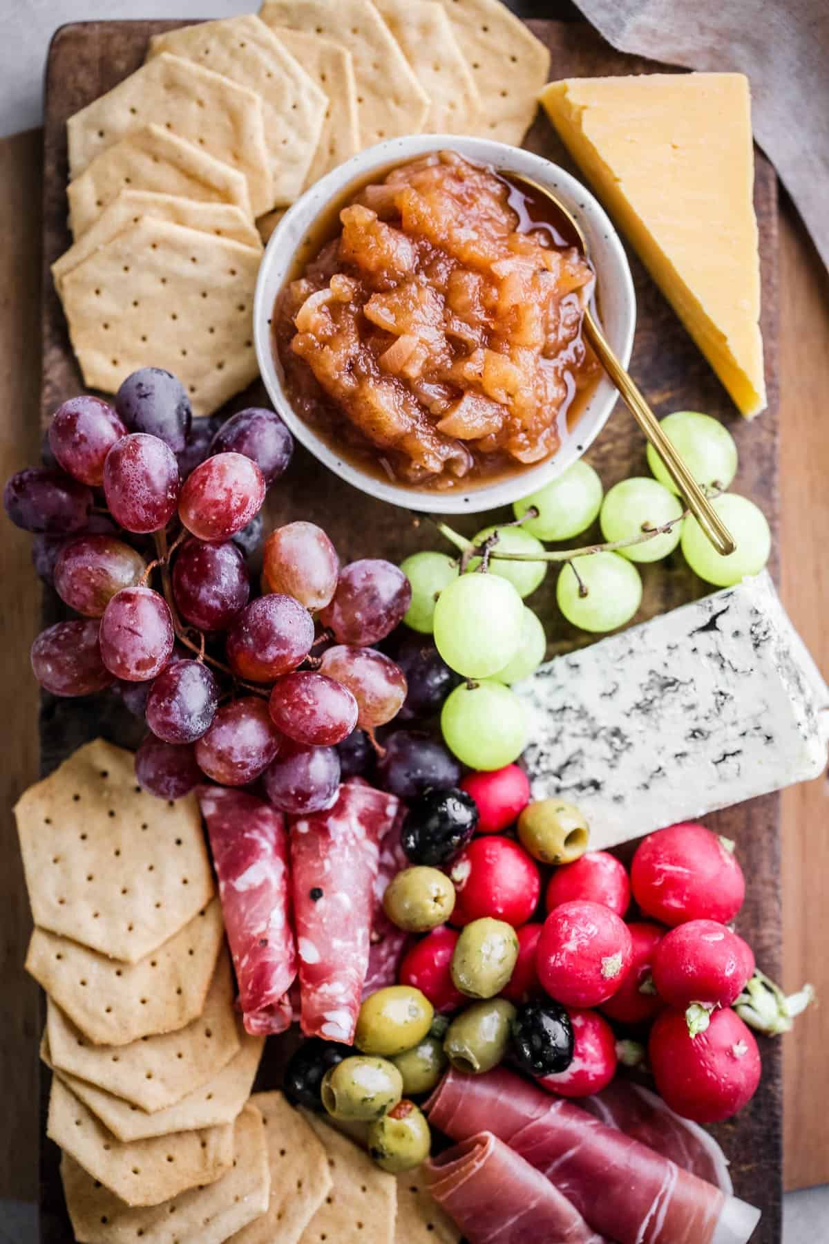 Bowl of Apple Ginger Chutney on a board surrounded by crackers, cheese, charcuterie, grapes.