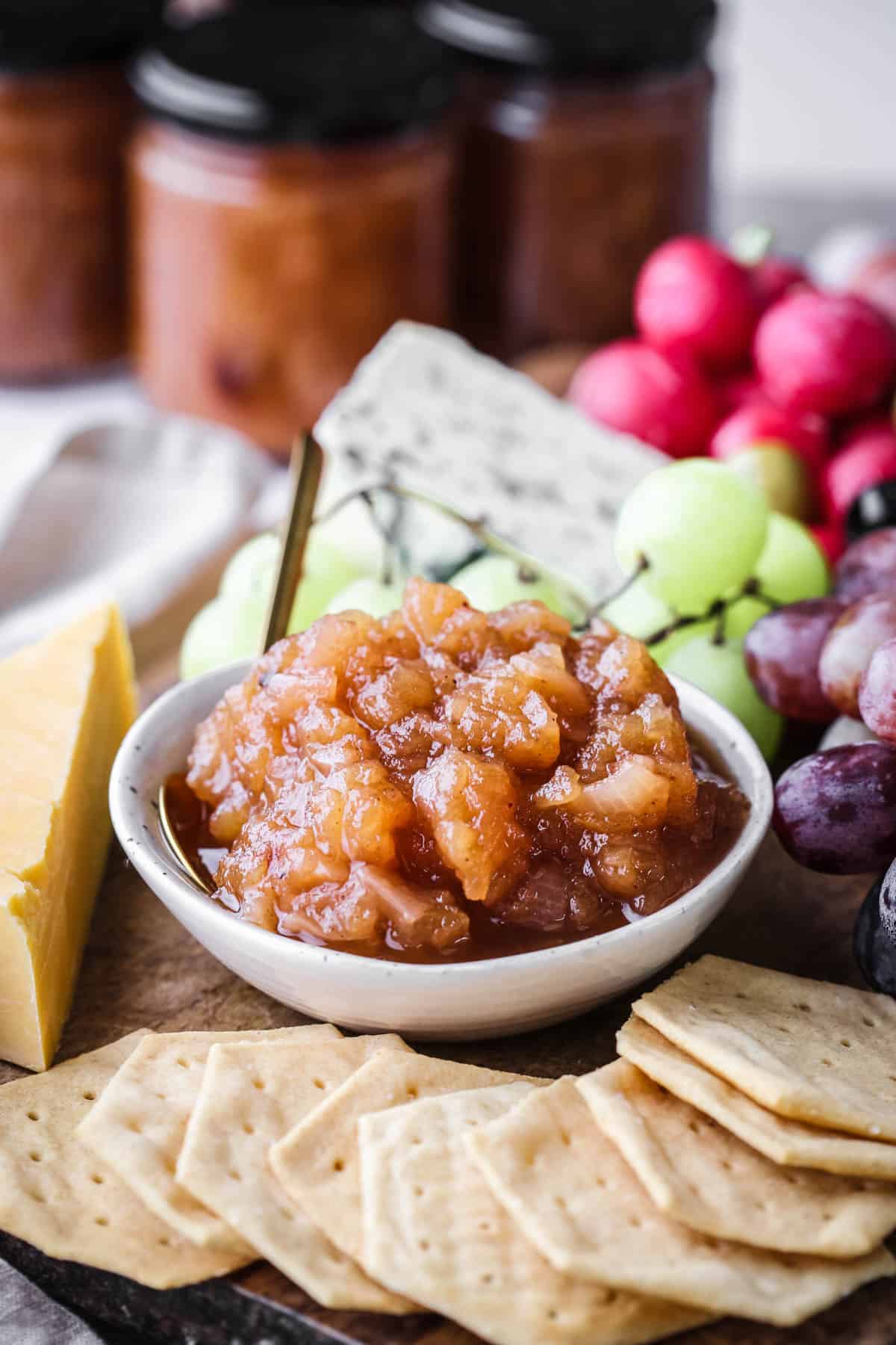 Bowl of Apple Ginger Chutney on a board surrounded by crackers, cheese, charcuterie, grapes.