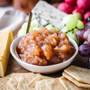 Bowl of Apple Ginger Chutney on a board surrounded by crackers, cheese, charcuterie, grapes.