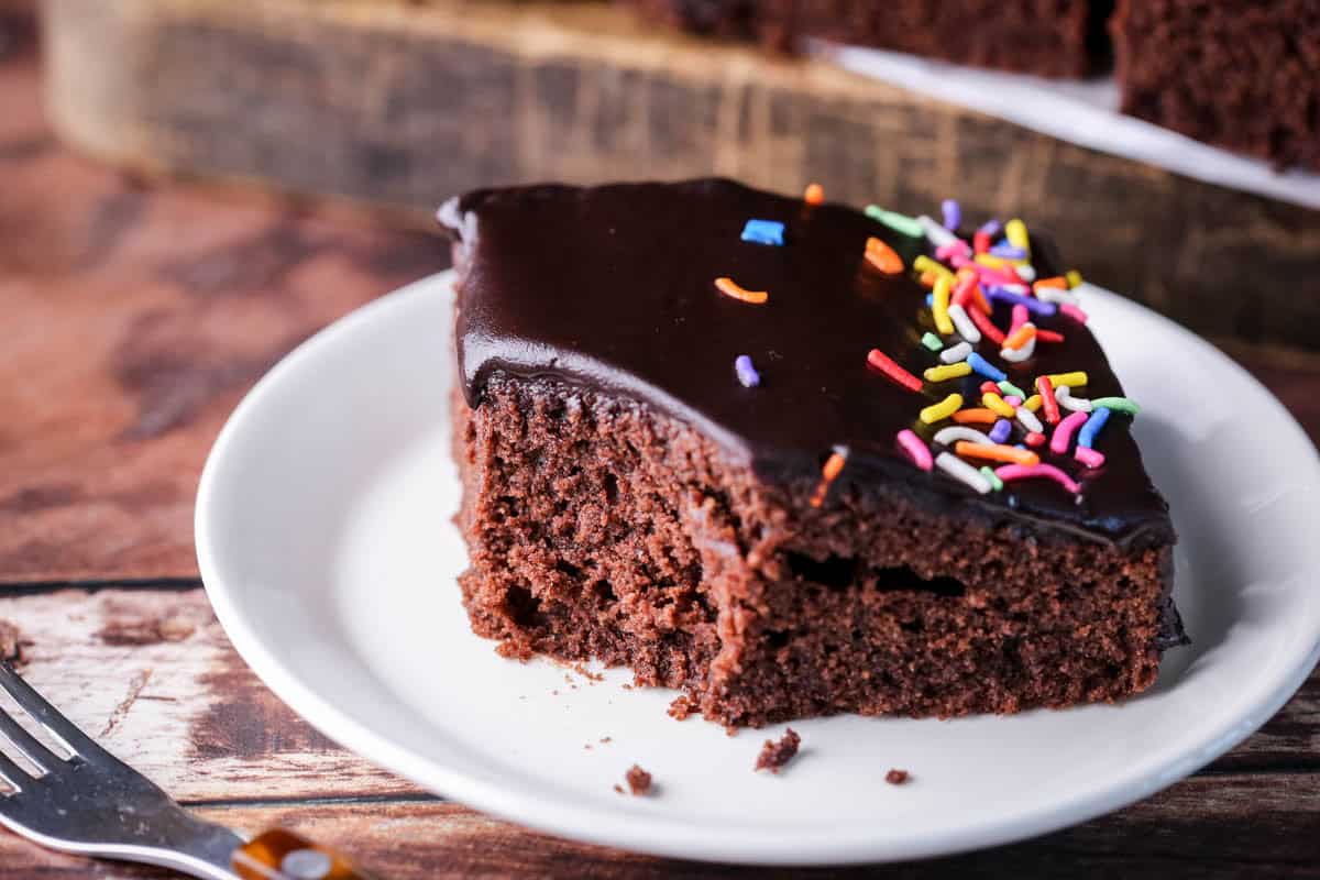 Chocolate School Cake topped with smooth chocolate icing and colourful sprinkles. With bite taken out showing soft and tender chocolate crumb.