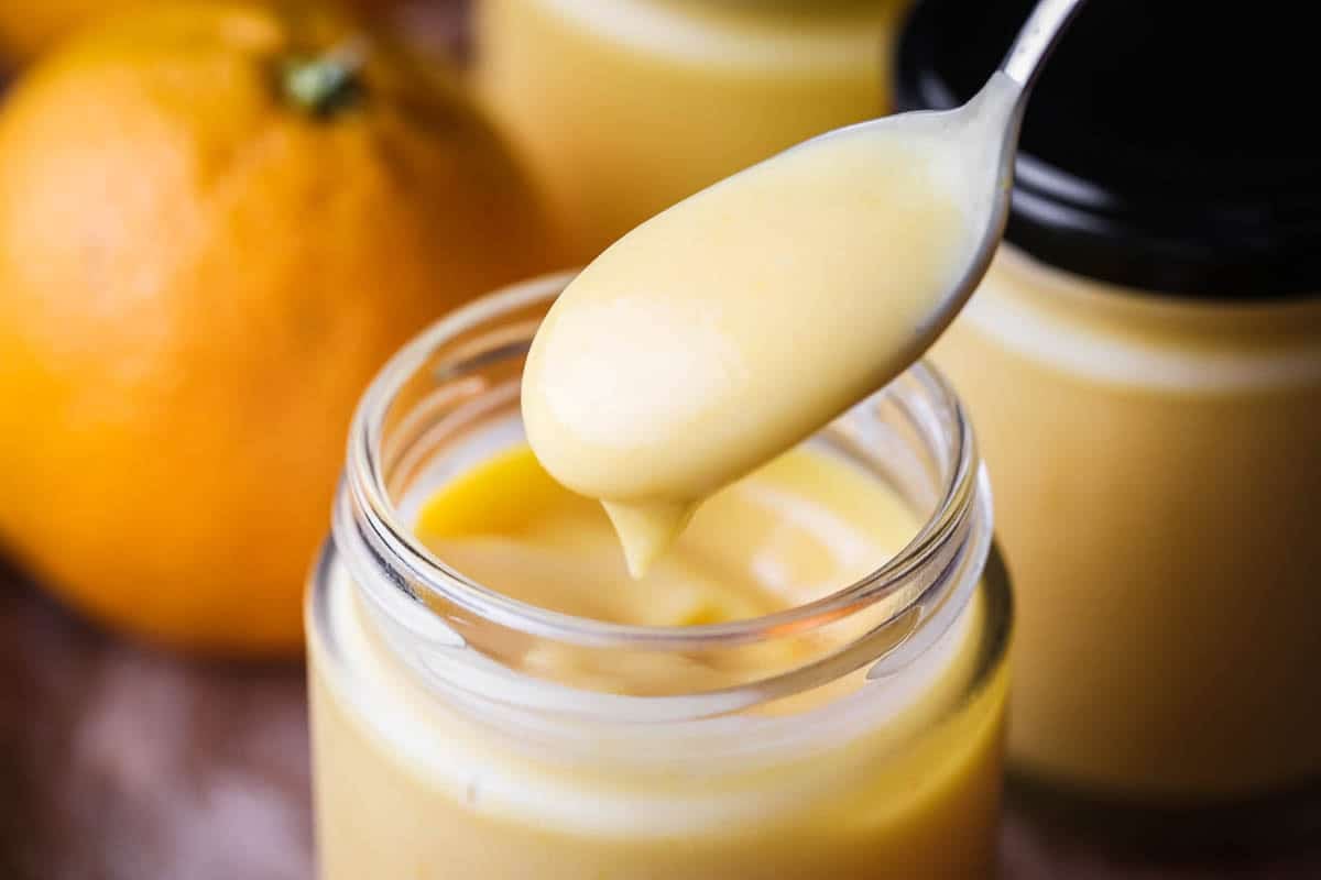 Spoon dipping into jar of freshly made orange curd showing thick ultra smooth preserve with a bright colour.