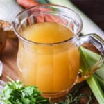 Homemade Chicken or Turkey Stock in a jug, a deep rich golden liquid, surrounded by vegetables and aromatics.