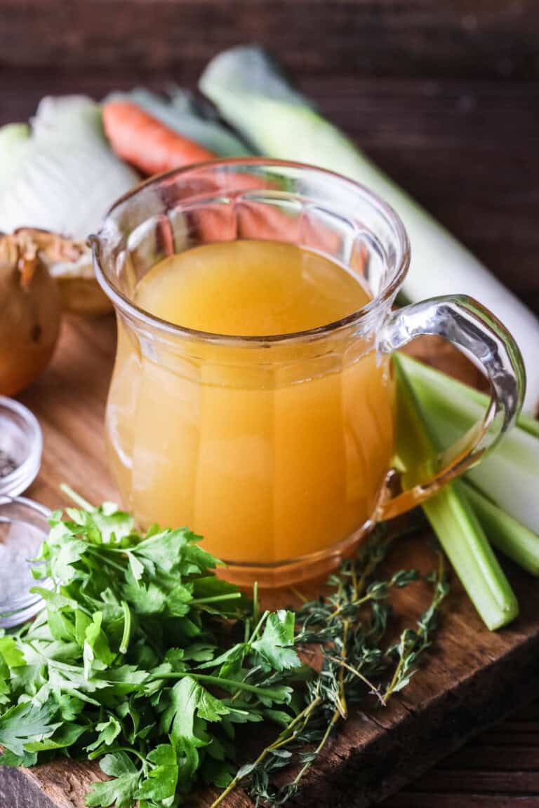 Homemade Chicken or Turkey Stock in a jug, a deep rich golden liquid, surrounded by vegetables and aromatics.