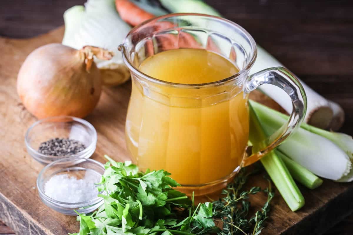 Homemade Chicken or Turkey Stock in a jug, a deep rich golden liquid, surrounded by vegetables and aromatics.