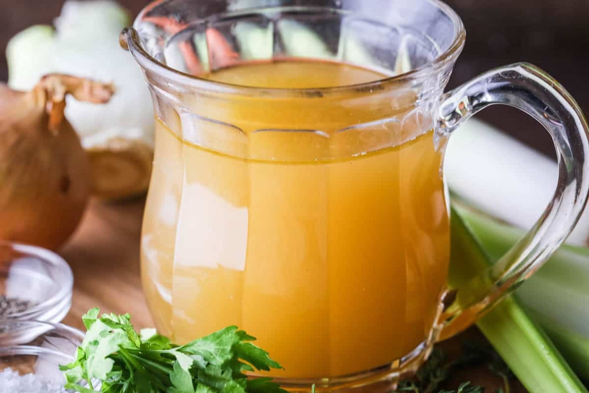 Homemade Chicken or Turkey Stock in a jug, a deep rich golden liquid, surrounded by vegetables and aromatics.