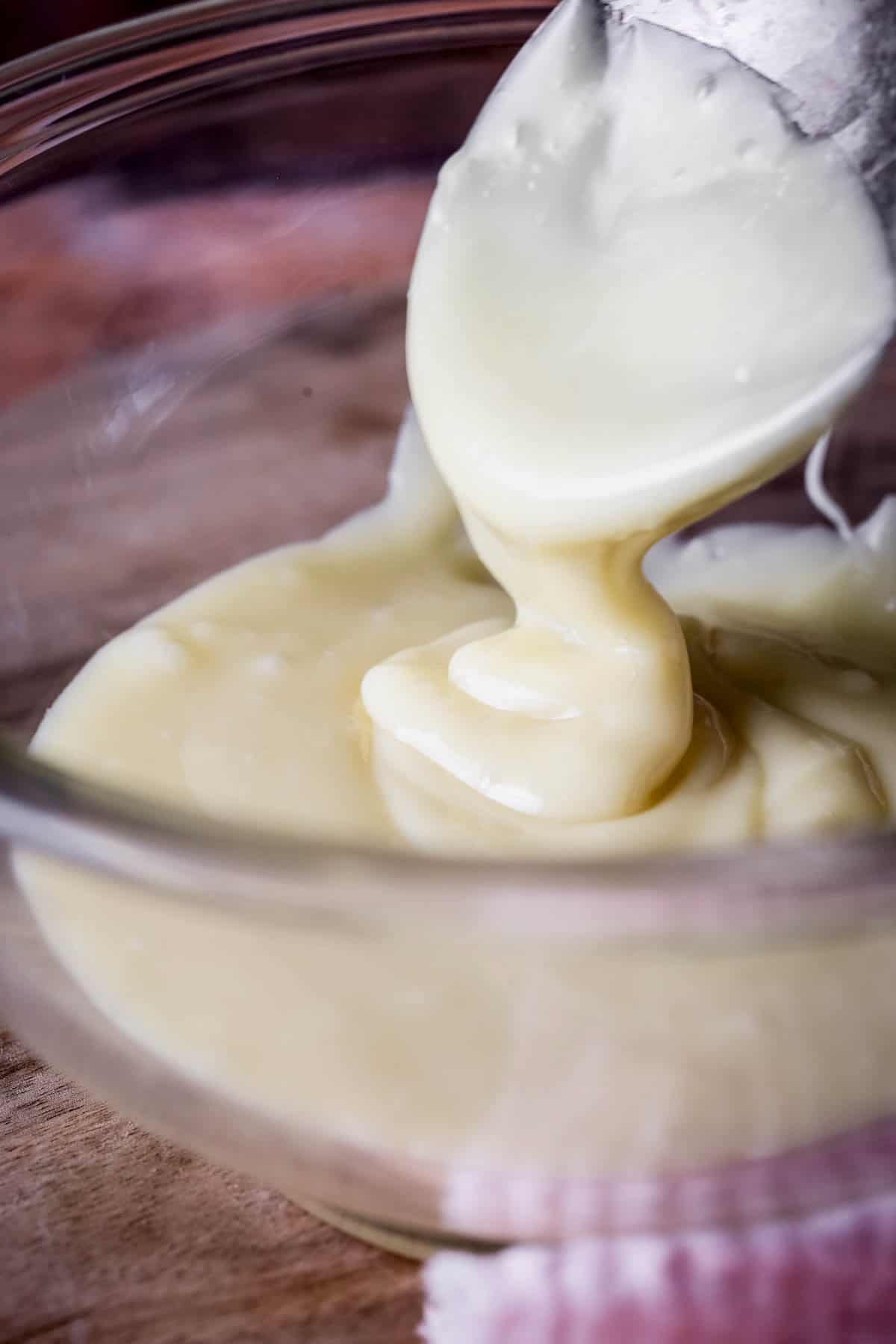 White Chocolate Ganache smooth and very pourable being lifted up by spoon in a bowl.