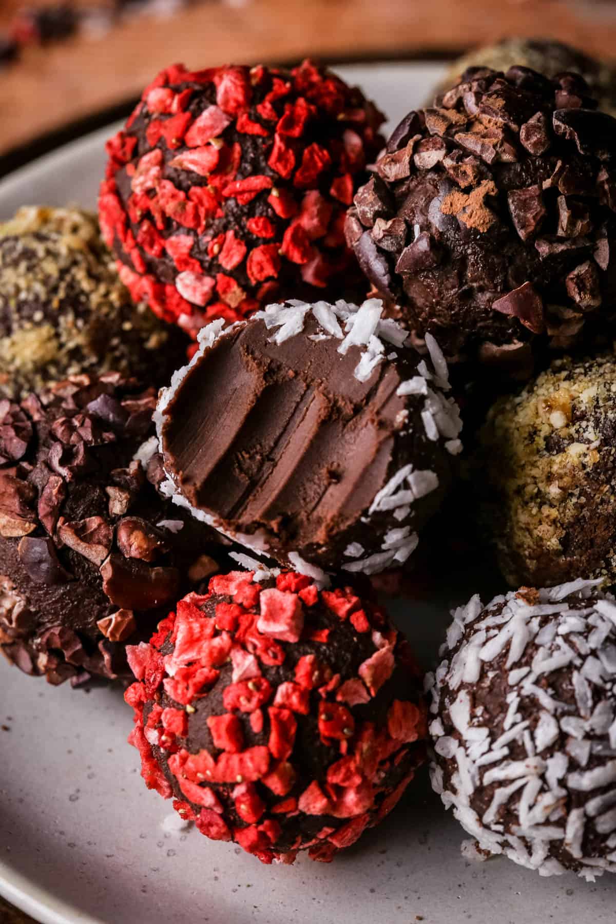 Pile of homemade chocolate truffles on a plate rolled in a variety of coatings. One with a bite taken out showing soft creamy centre.