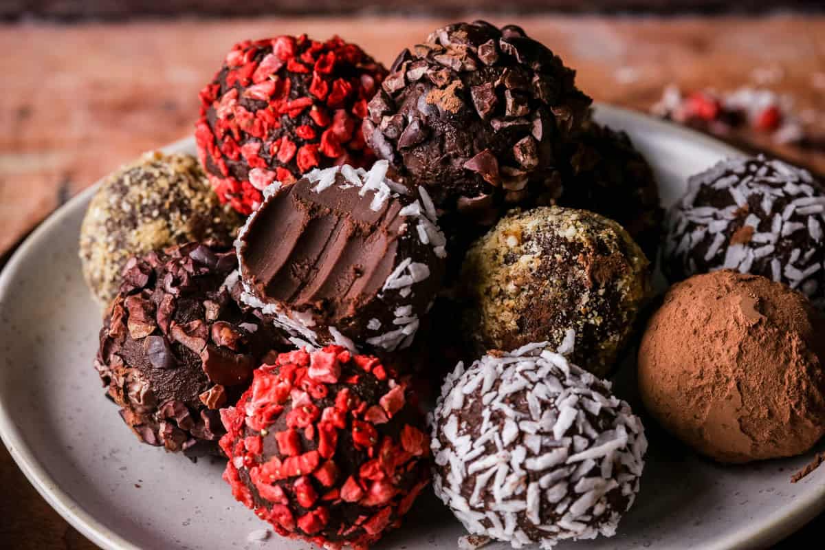 Pile of homemade chocolate truffles on a plate rolled in a variety of coatings. One with a bite taken out showing soft creamy centre.