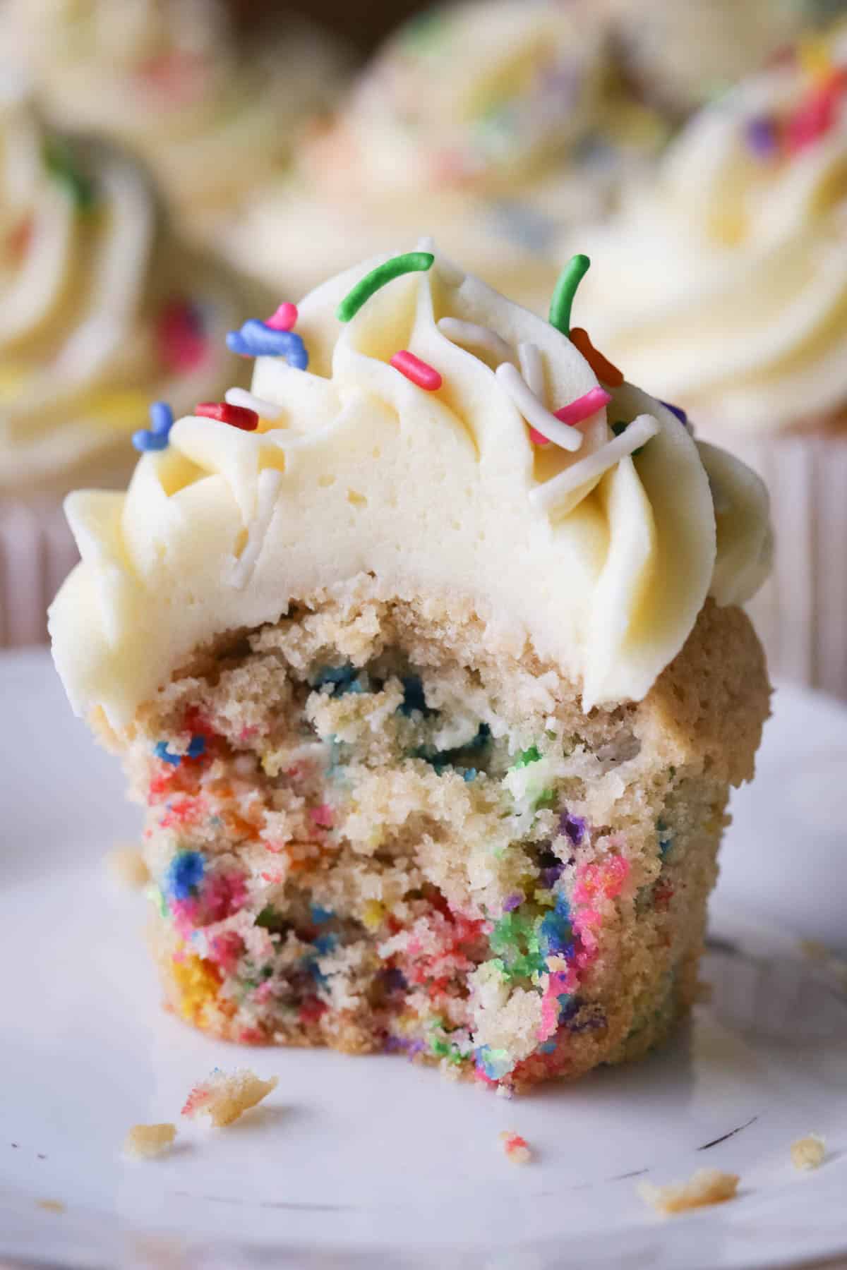 Gluten-Free Funfetti Cupcake on a plate with a bite taken out showing the soft tender fluffy crumb which is packed with colourful sprinkles.