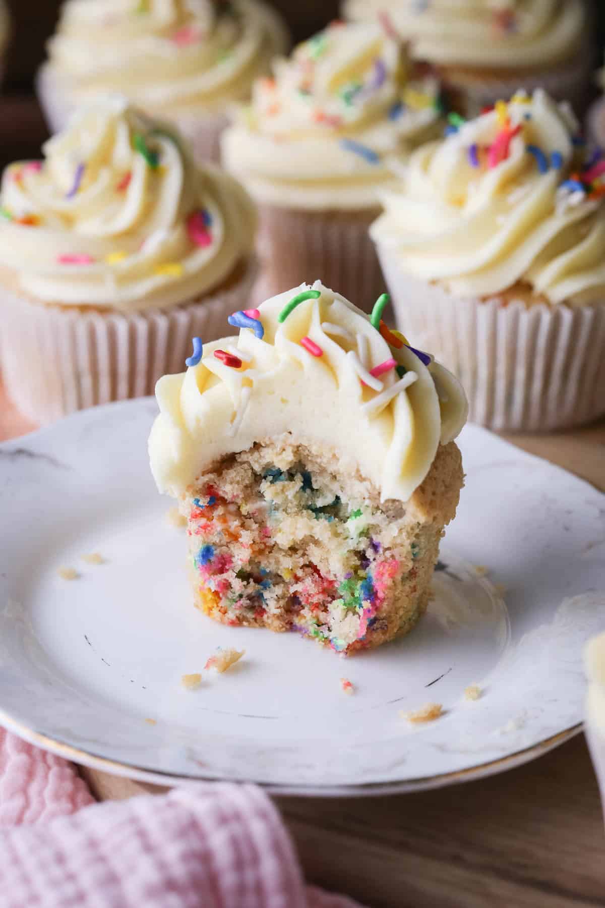 Gluten-Free Funfetti Cupcake on a plate with a bite taken out showing the soft tender fluffy crumb which is packed with colourful sprinkles.