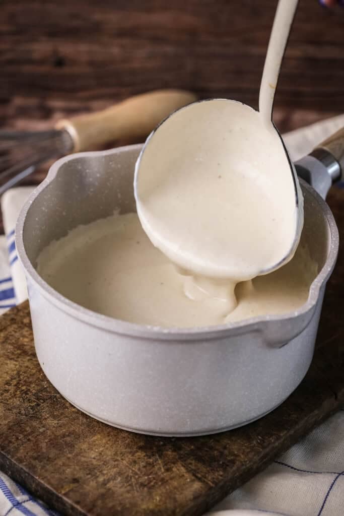 Béchamel in a saucepan with large ladle scooping some up showing how lusciously thick and creamy it is.