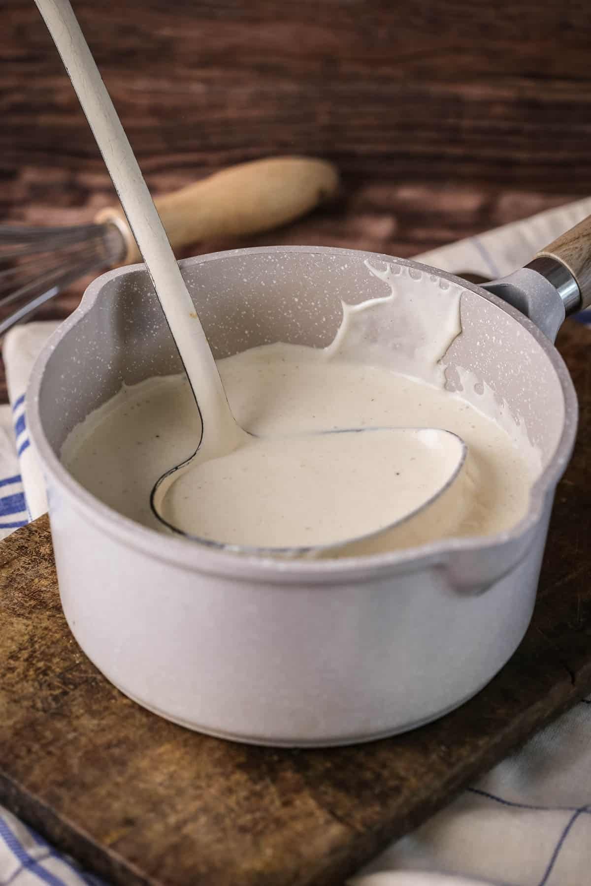 Béchamel in a saucepan with large ladle scooping some up showing how lusciously thick and creamy it is.