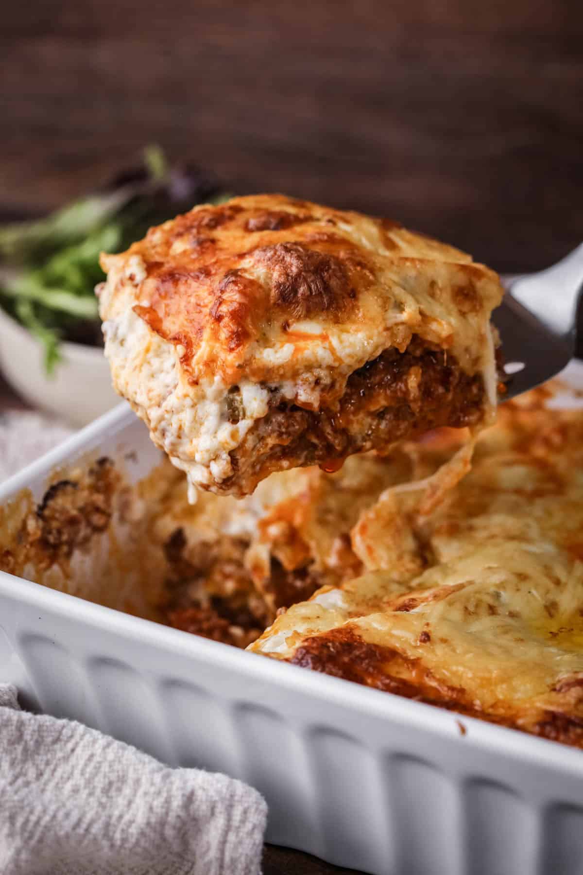 Slice of lasagne lifted out of baking dish, showing cheesy meaty middle.