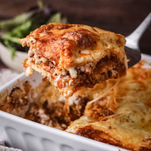 Slice of lasagne lifted out of baking dish, showing cheesy meaty middle.