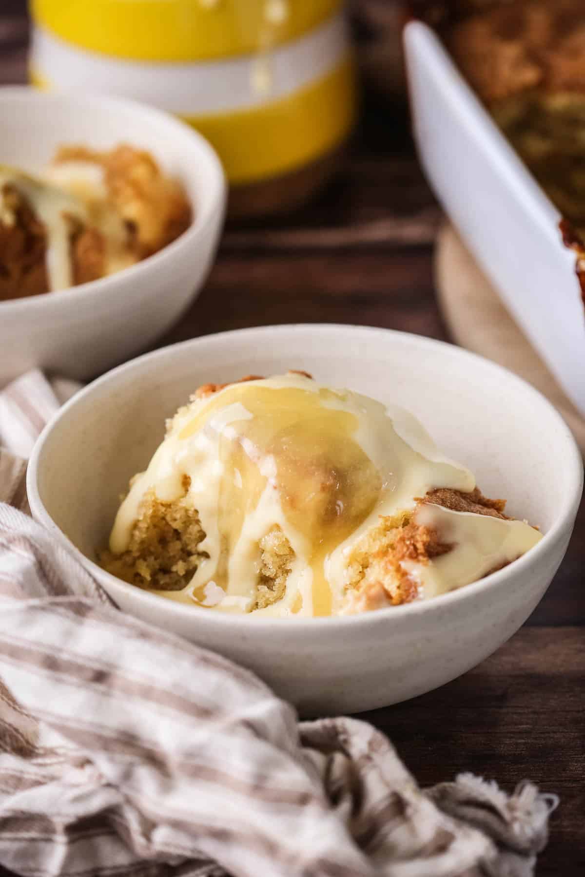 Portion of lemon pudding in a bowl with custard poured over top and extra lemon curd drizzled over.
