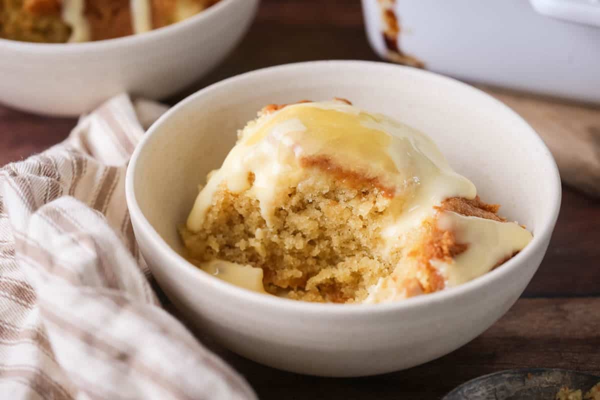 Portion of lemon pudding in a bowl with custard poured over top and extra lemon curd drizzled over.