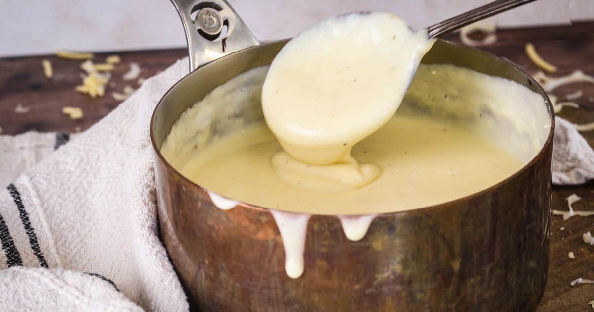 Spooning gluten-free cheese sauce from saucepan showing velvety creamy texture.