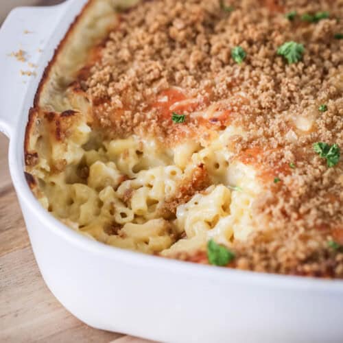 A portion of mac and cheese has been removed from serving dish showing ultra creamy and cheesy sauce clinging to tender macaroni pasta. With a golden crisp topping and fresh parsley.
