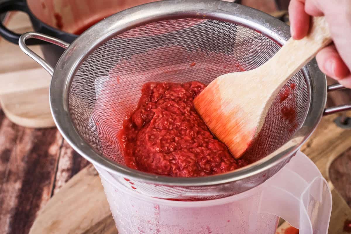 Chinois full of raspberry pulp and wooden spoon pushing it through into a measuring jug.