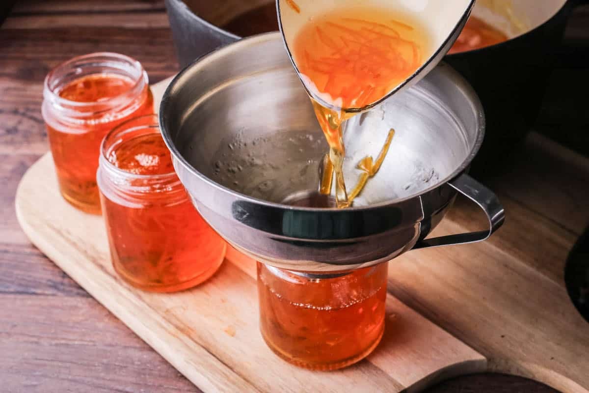 Ladle pouring out orange marmalade into jars through a jam funnel.