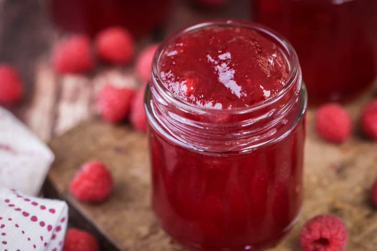 Pot of Seedless Raspberry Jam surrounded by raspberries on a wooden board.